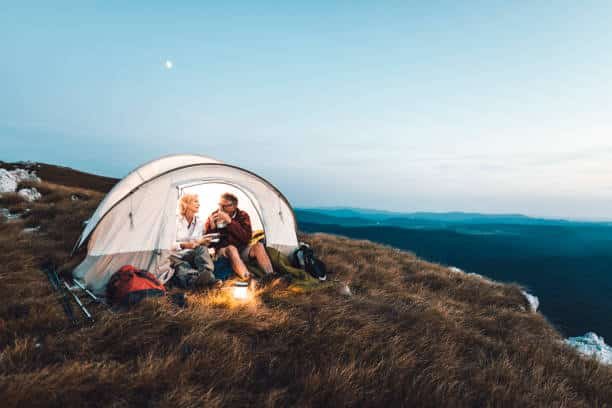 A couple sitting inside a camping tent in the mountains, enjoying the evening together with a lantern, surrounded by a vast landscape of rolling hills at sunset.