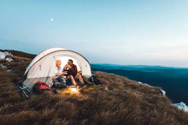 A couple sitting inside a camping tent in the mountains, enjoying the evening together with a lantern, surrounded by a vast landscape of rolling hills at sunset.