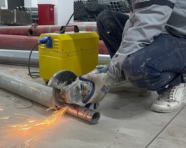 A worker using a portable grinder to cut a metal pipe on a construction site, with sparks flying from the cutting action and a nearby portable power supply in the background.