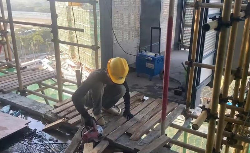 A construction worker wearing safety gear, operating a power tool on a scaffolding platform, with a portable power source visible in the background, in a high-rise construction setting.