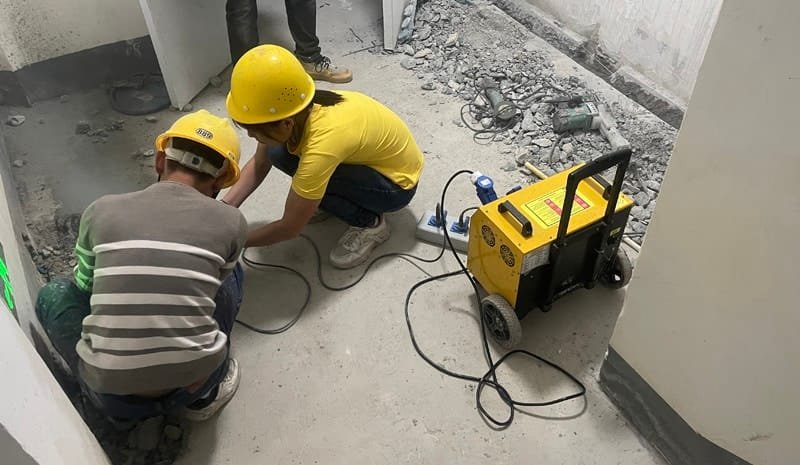 Two workers in safety helmets using power tools on a construction site, with a portable generator providing energy in the background, amidst scattered debris.